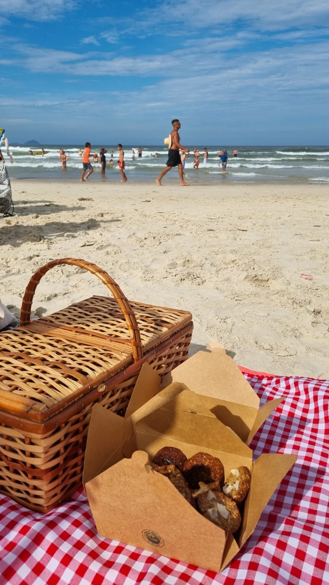Wicker basket and a box of snacks on a checkered cloth at the beach.