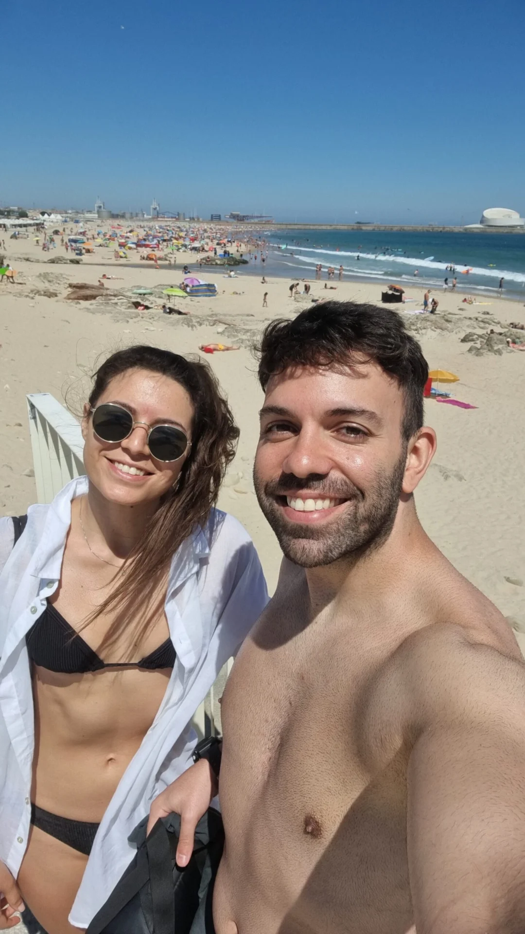 A smiling couple taking a selfie at a busy beach on a sunny day.