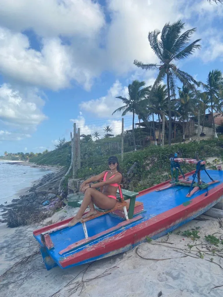 Woman sitting on a painted boat beside a tropical beach with palm trees and waves.