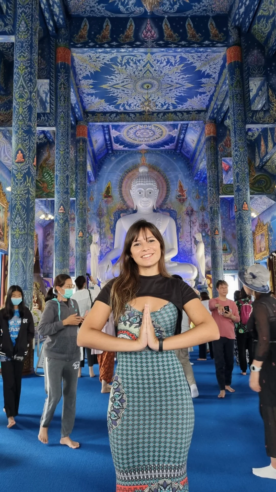 Woman posing with hands together inside a blue temple hall with a white Buddha statue behind her.