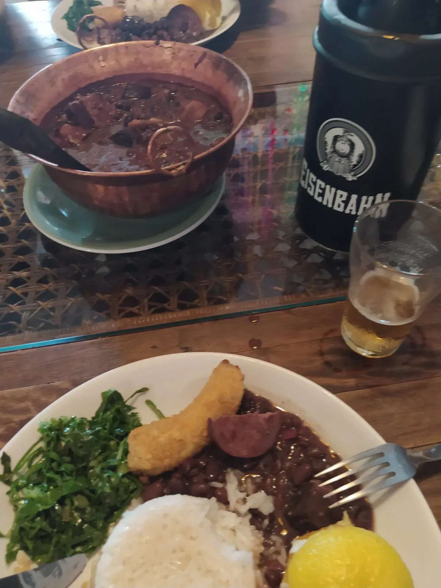 Plate of Brazilian rice, beans, greens, and fried sides beside a drink on a table.