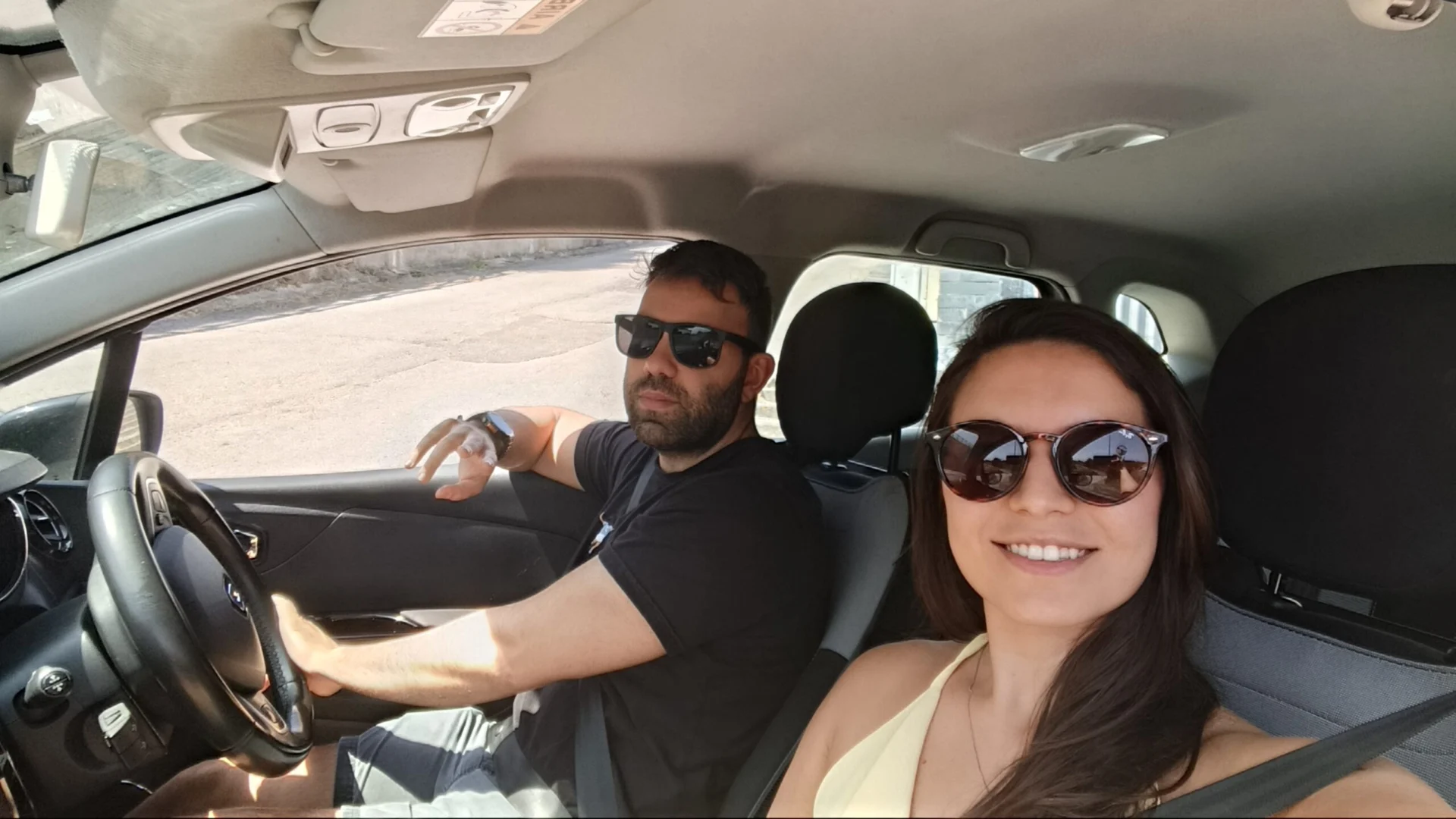 Two people sitting in a car, with the driver at the wheel and a passenger taking a selfie.