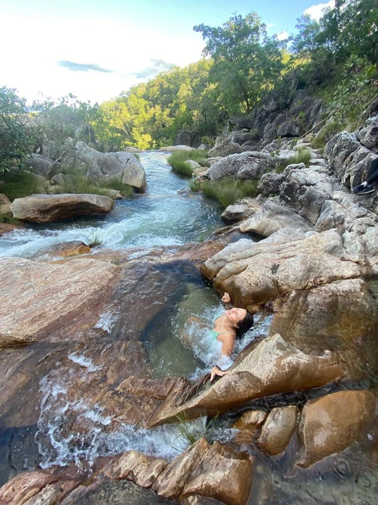 A person relaxes in a shallow rocky river pool with clear water and trees in the background.