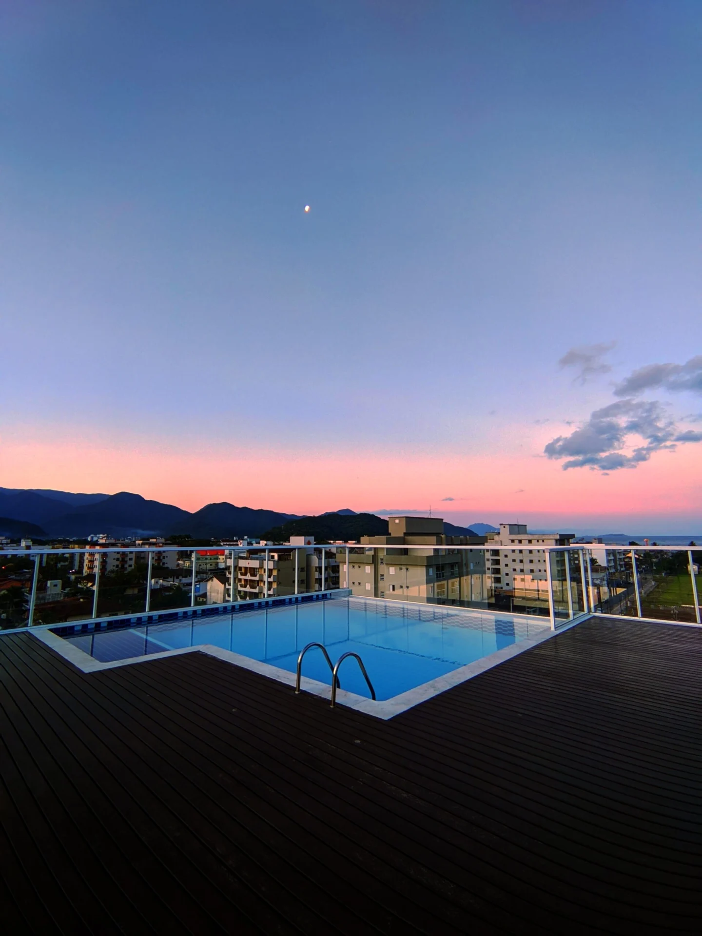 A rooftop pool overlooks city buildings and distant mountains under a pink and blue sunset sky.