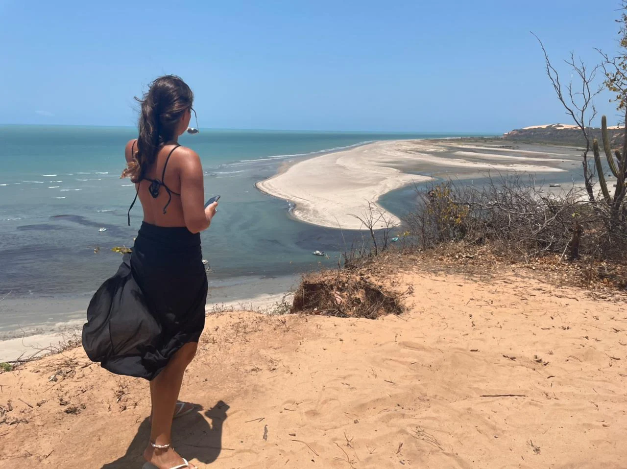 Woman standing on a sandy cliff overlooking a wide blue coastline with a pale sandbar curving into the sea.