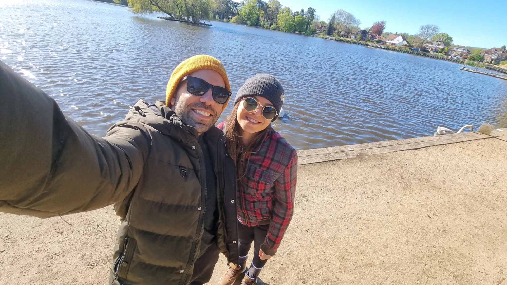 A smiling couple taking a selfie beside a lake on a sunny day.