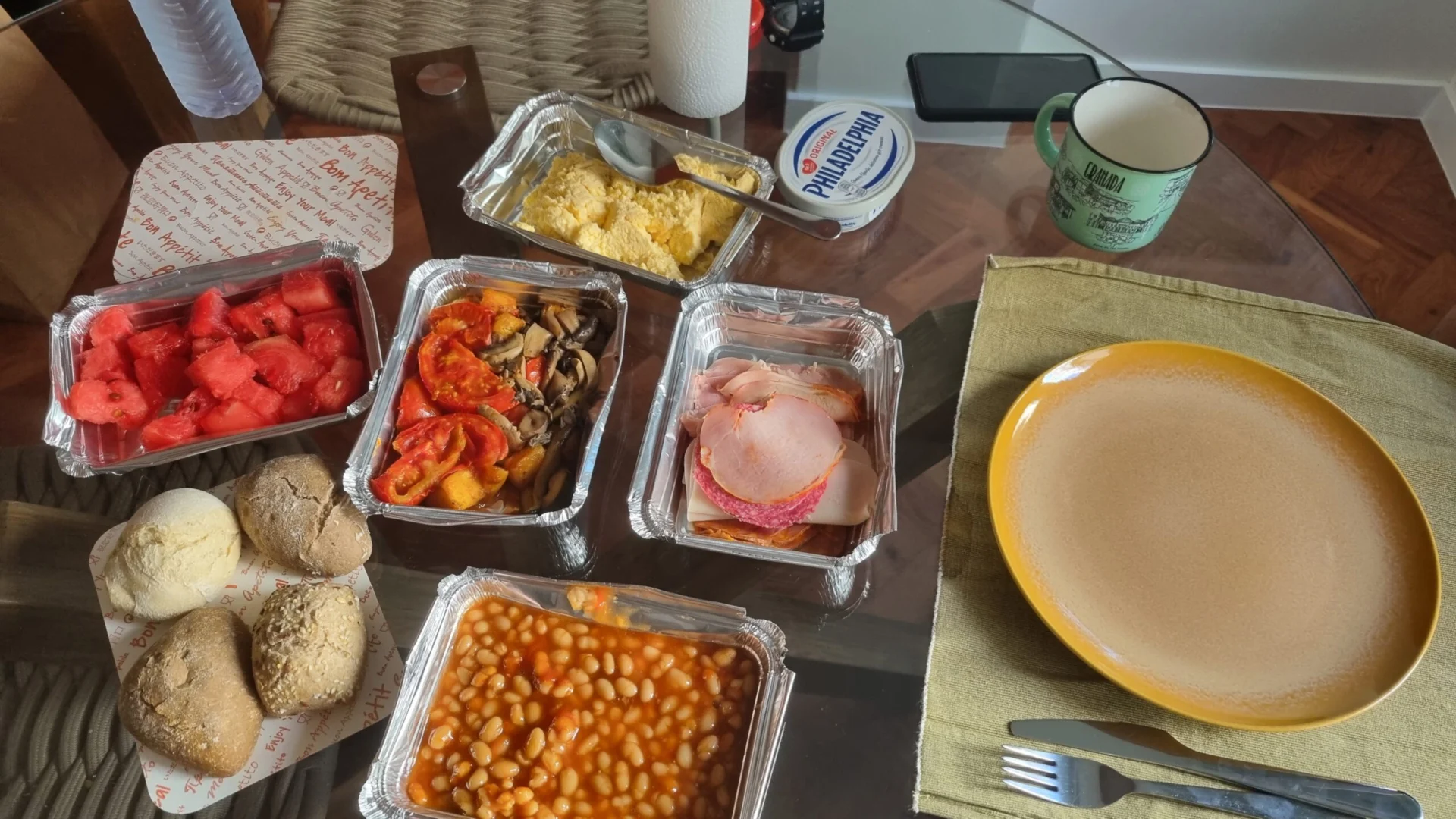 A table holds packaged meals, fruit, bread rolls, and other food items.