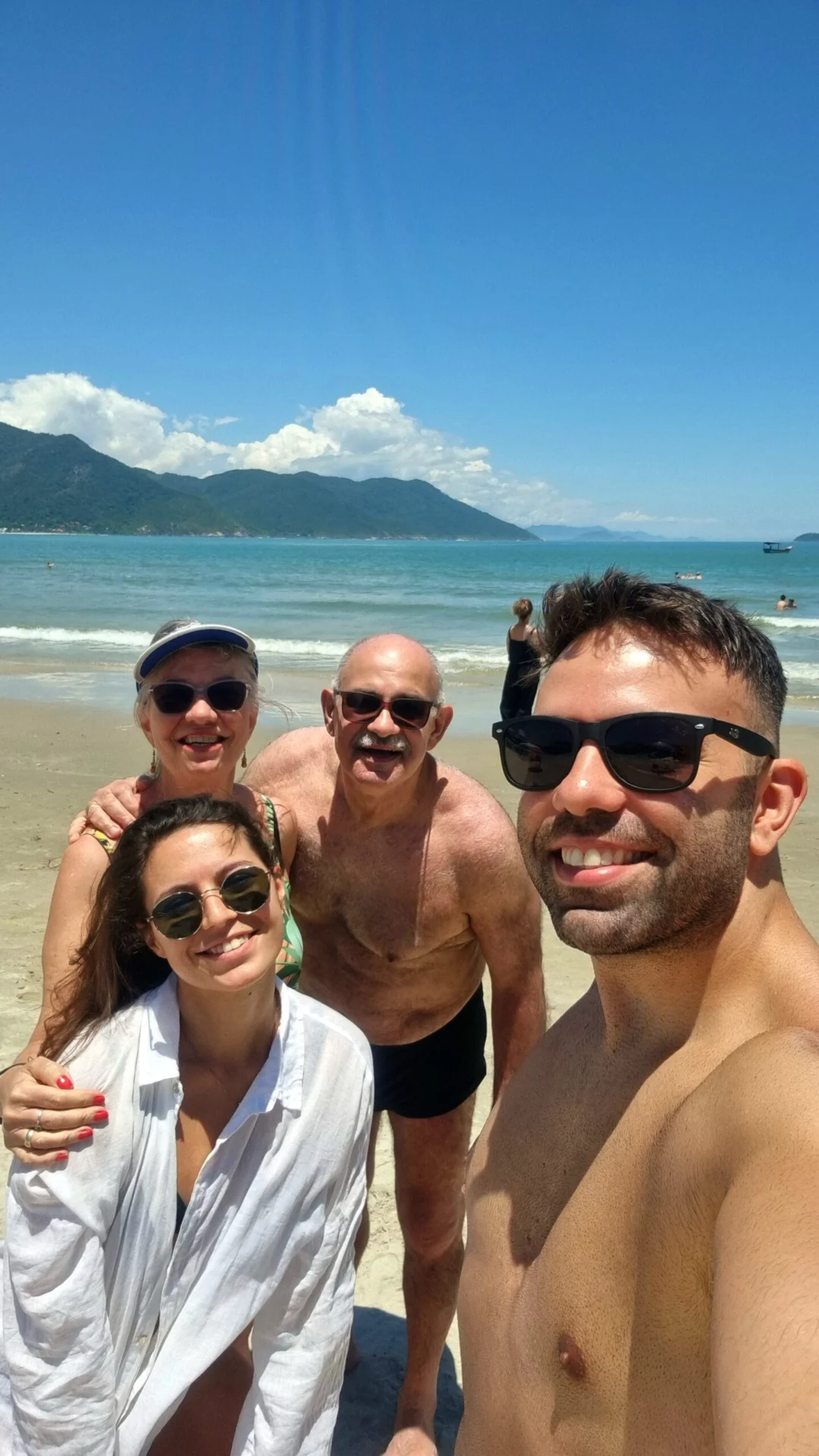 Four people smiling for a selfie on a sunny beach with sea and mountains behind them.