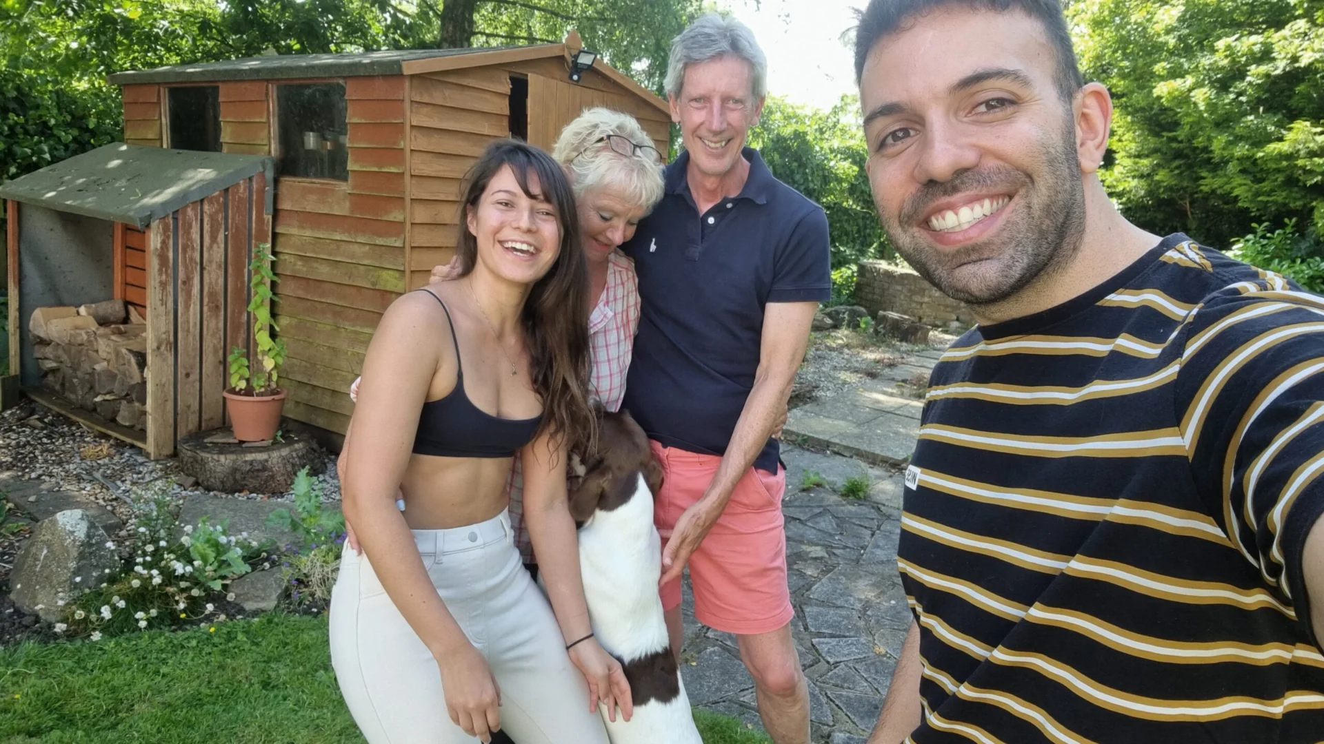 Group of four smiling people standing outside a small wooden cottage with a dog in front.