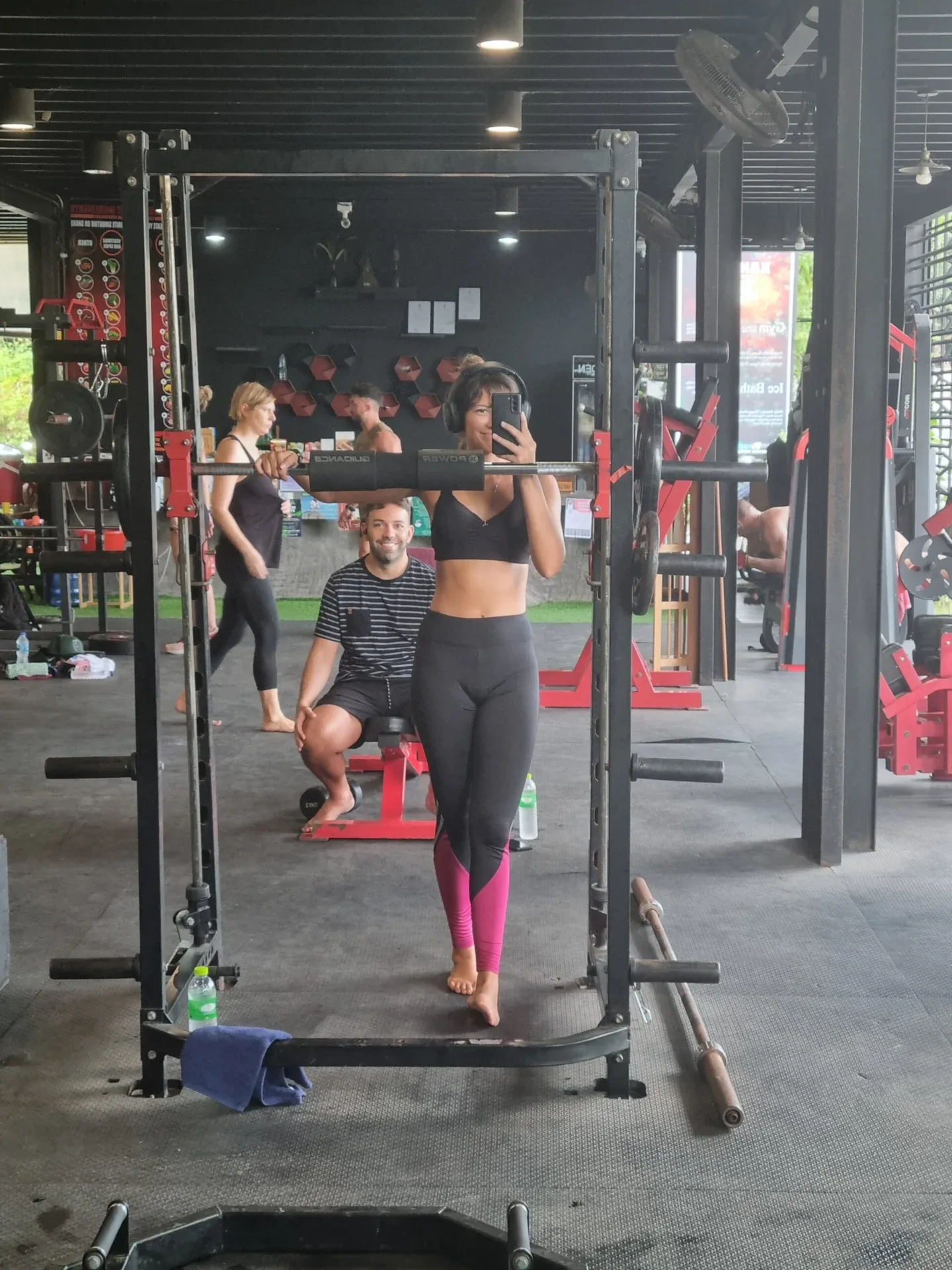 A woman takes a mirror selfie in a gym while people work out behind her on weight machines and benches.