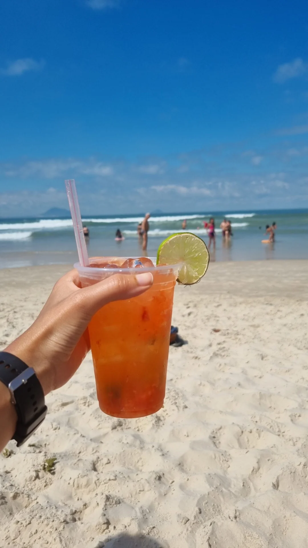 Hand holding a cold orange drink with a lime slice on a sunny beach.