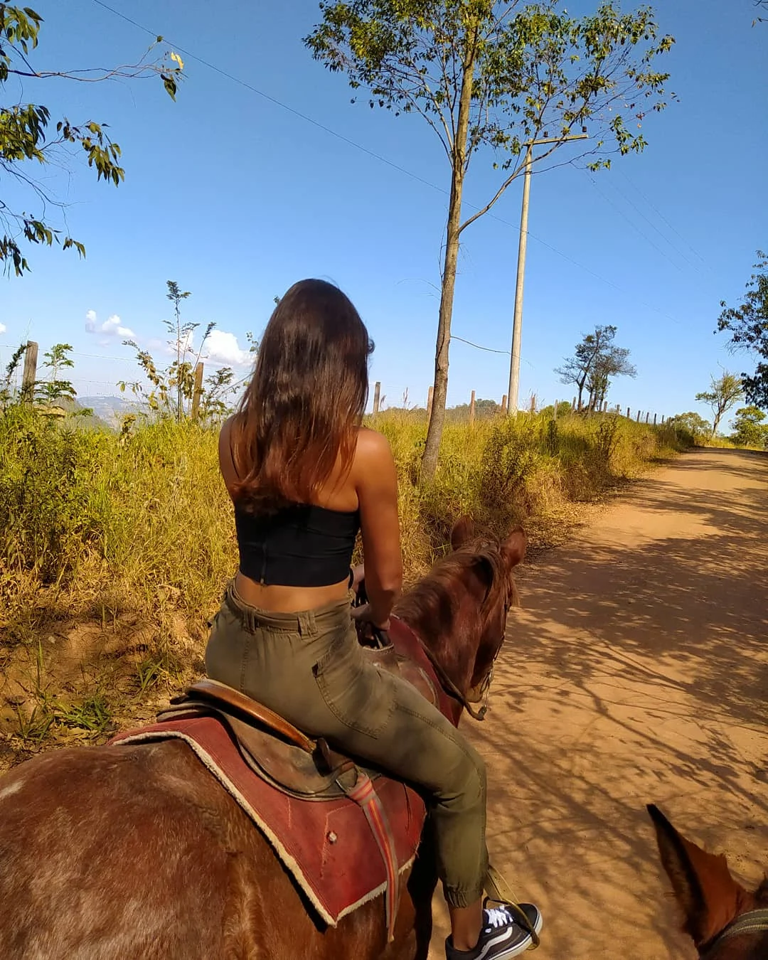 Woman riding a horse on a dirt trail through dry countryside with trees and open sky.