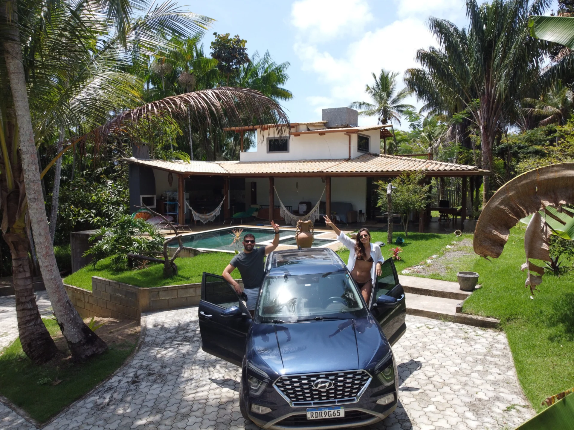 Two travelers stand beside a car in front of a tropical house with a pool and palm trees.