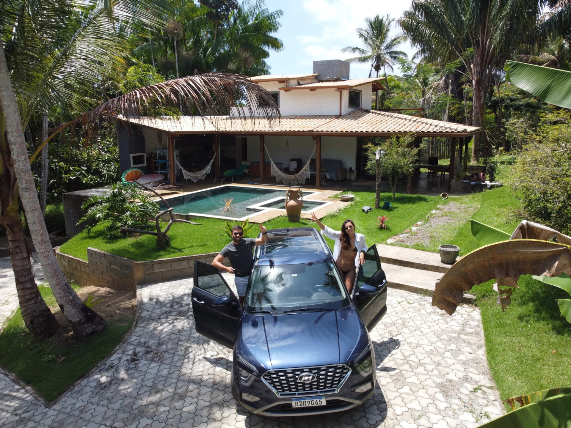 Two people stand by an open car door outside a tropical house with a pool and palm trees.