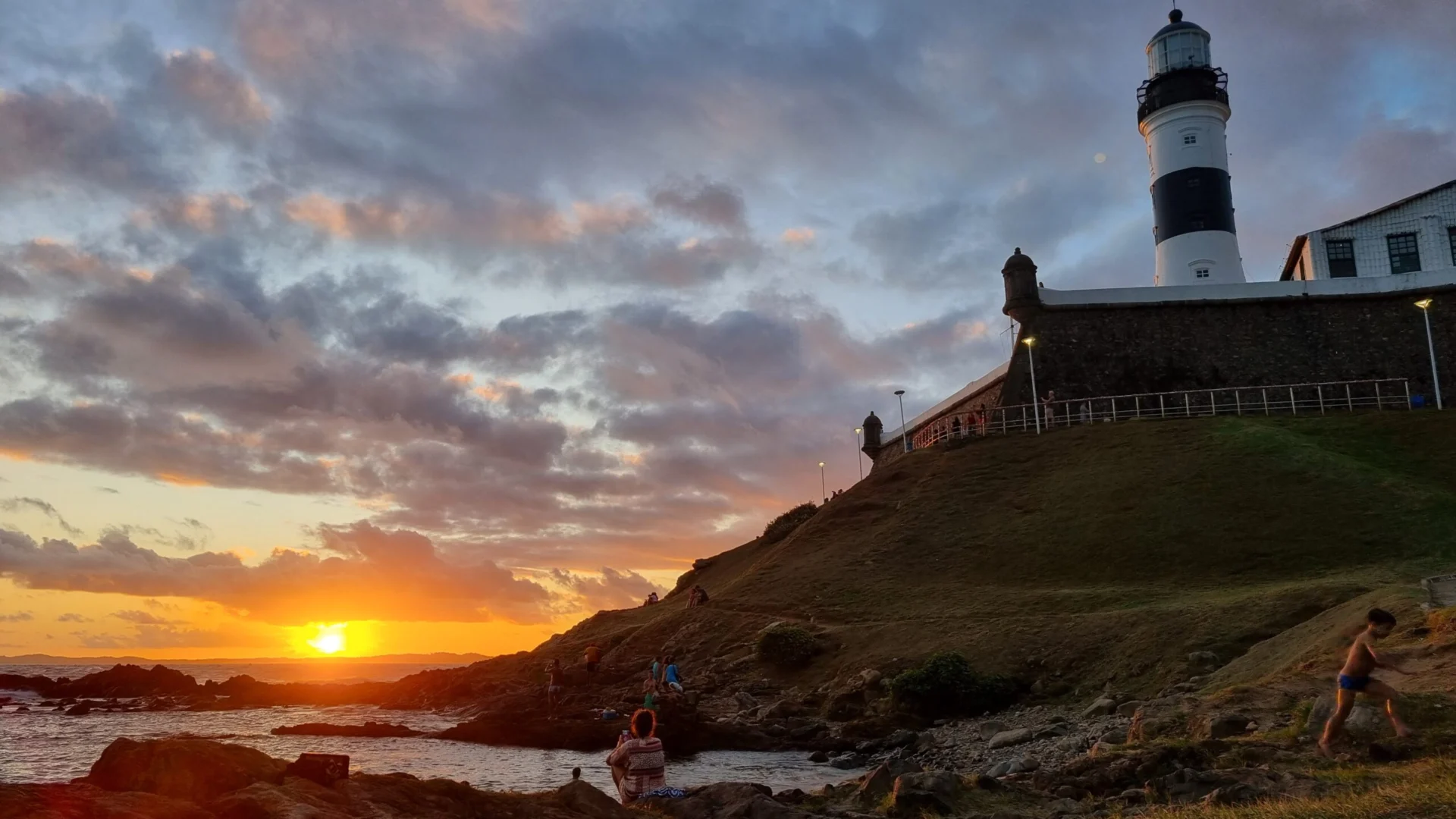 Lighthouse on a rocky coastline at sunset with colorful clouds over the sea.