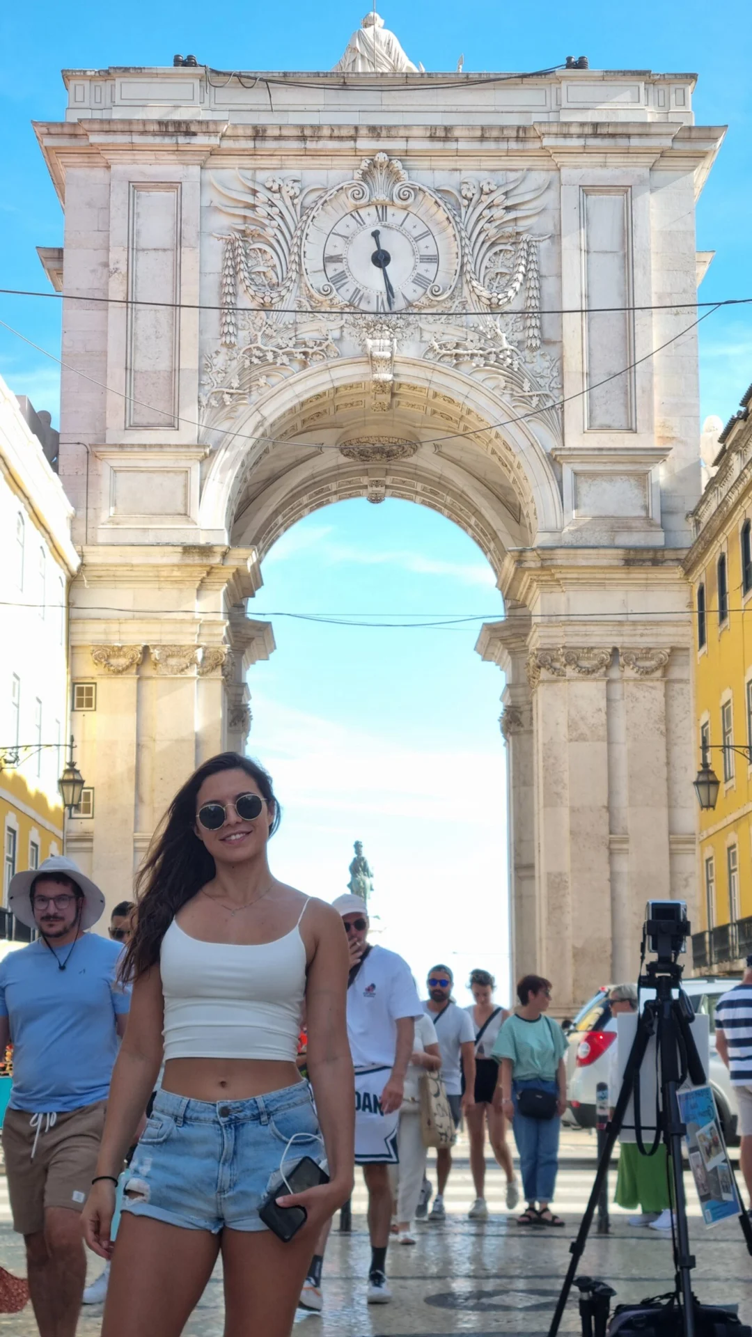 Woman standing in front of a large stone arch on a busy pedestrian street.