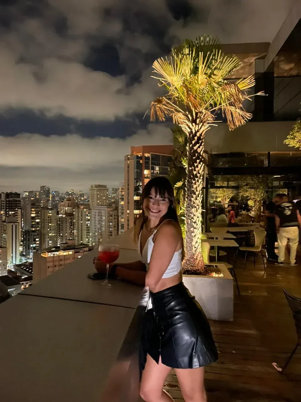 Woman smiling on a rooftop terrace with city lights and a palm tree at night.
