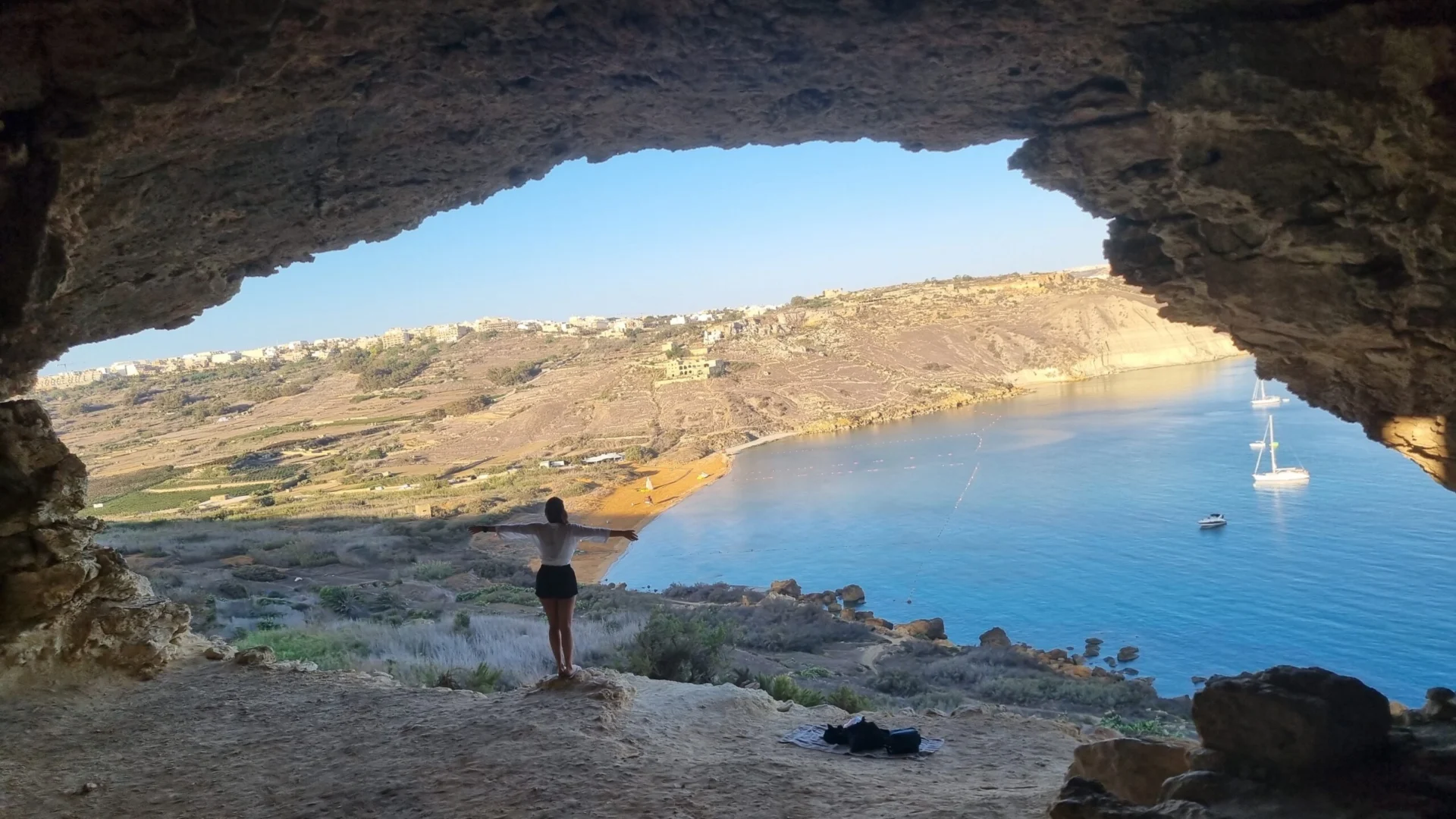 Person standing inside a rocky cave opening and looking out over a bay with clear blue water.