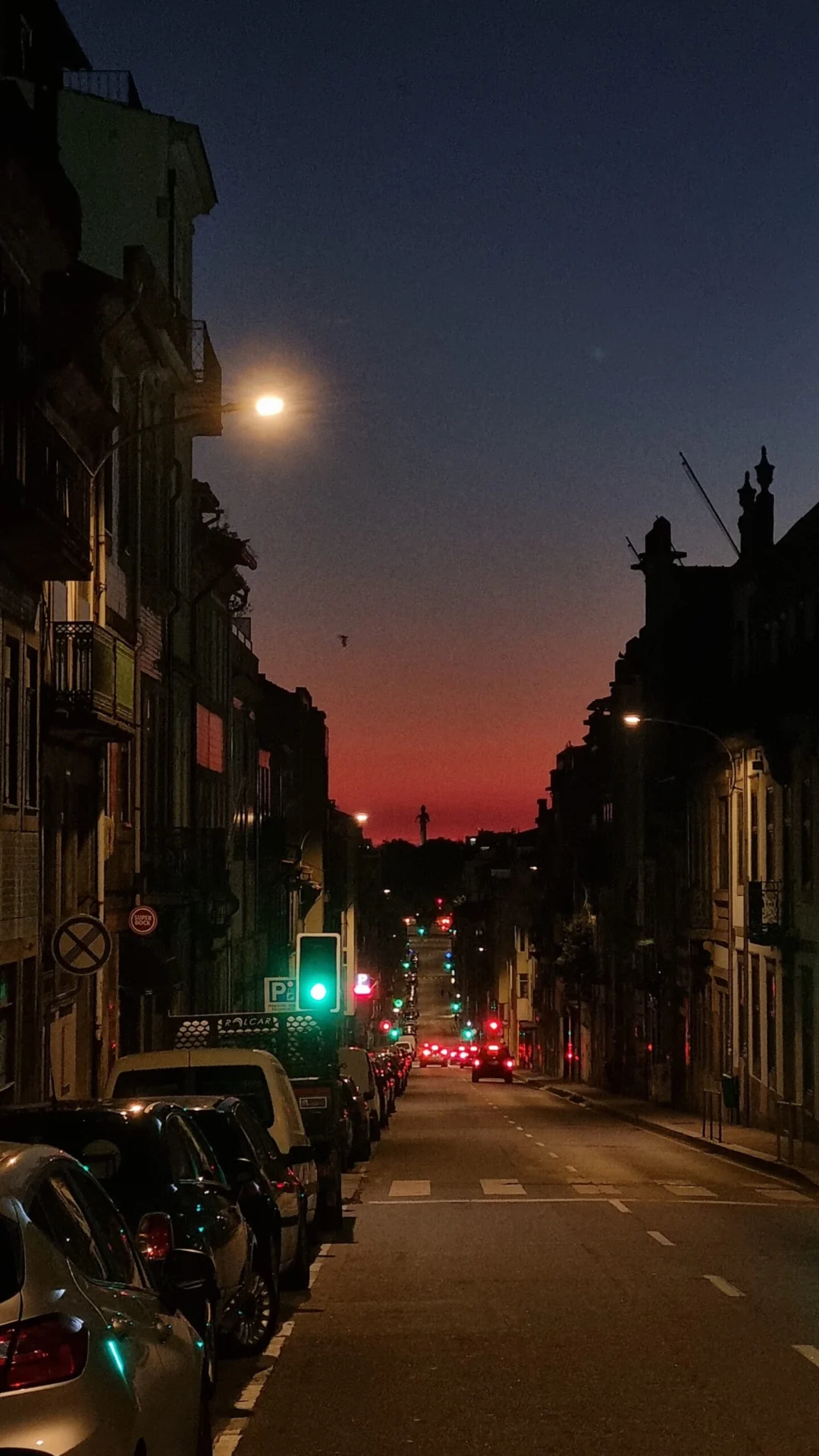 A narrow city street lined with parked cars and old buildings at dusk under a red and blue sky.