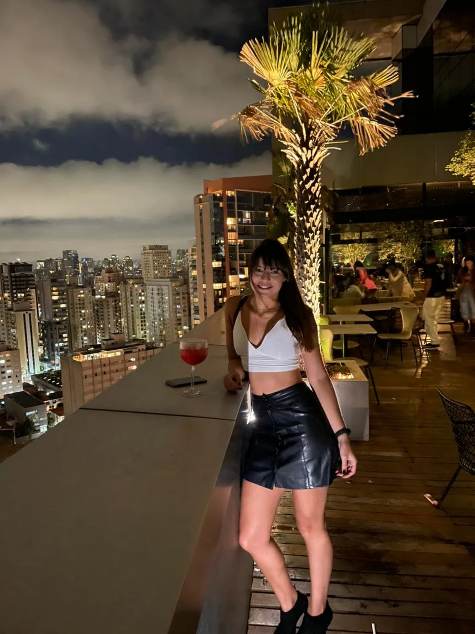 Woman standing on a rooftop terrace overlooking a city skyline at night with a palm tree lit beside her.