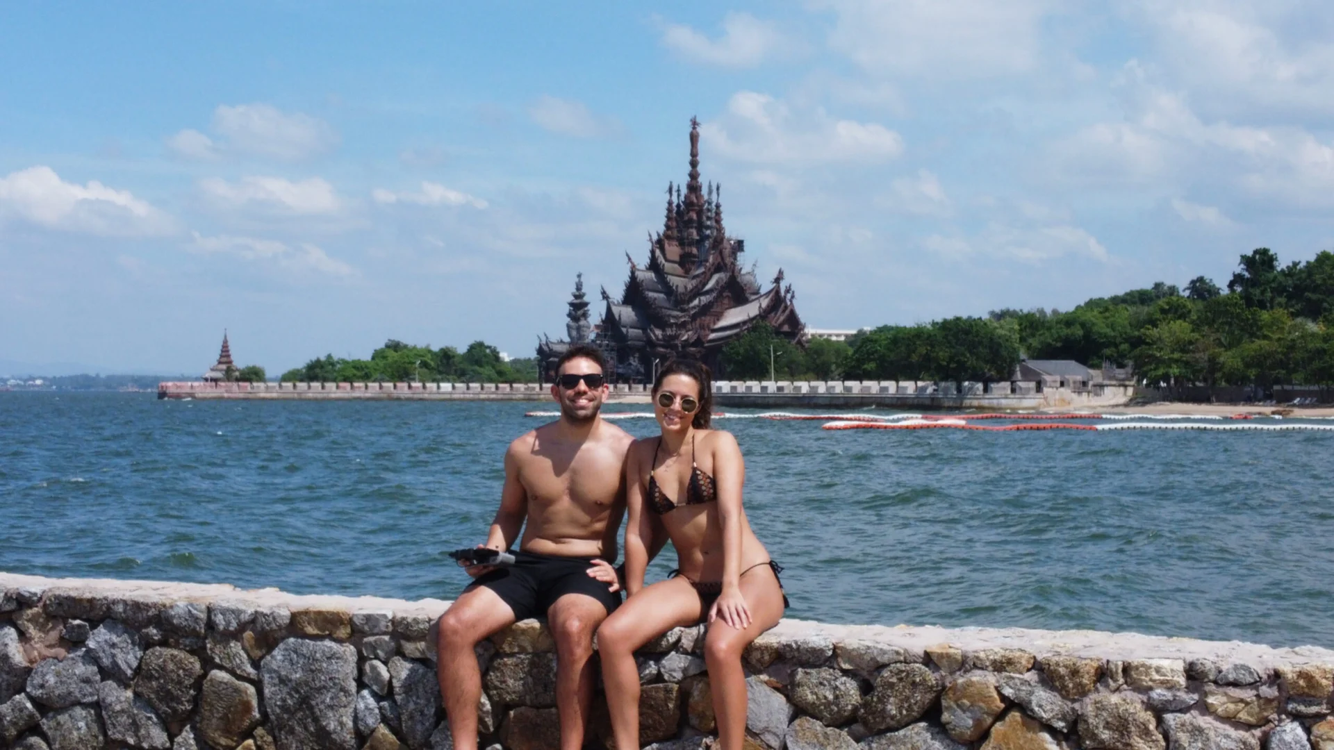 Two people sitting on a stone wall with the Sanctuary of Truth in the background by the water.