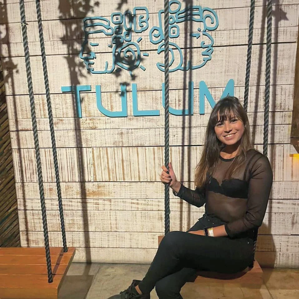 Woman posing indoors beside a suspended swing and a wall with the word Tulum.