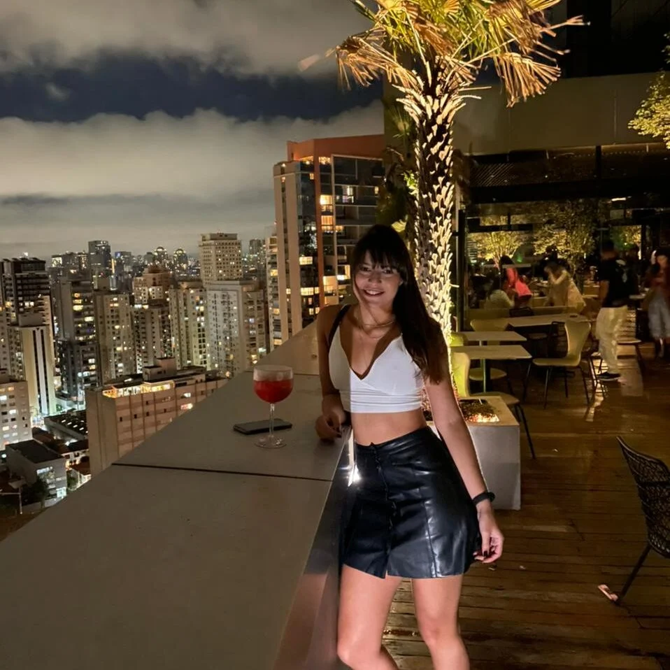 Woman standing on a rooftop at night with a city skyline and dining area behind her.
