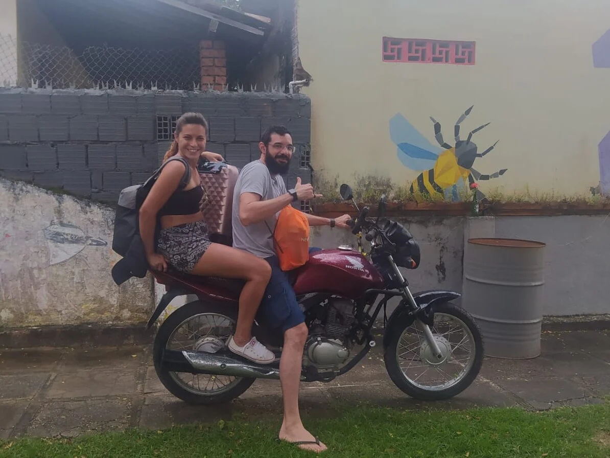 Two travelers riding together on a motorbike past a wall with a painted bee mural.