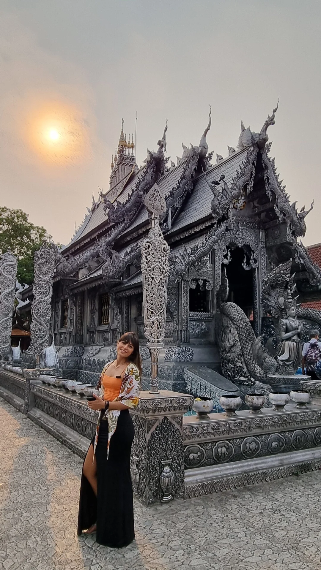 A woman stands beside the detailed silver exterior of Wat Si Suphan at sunset.