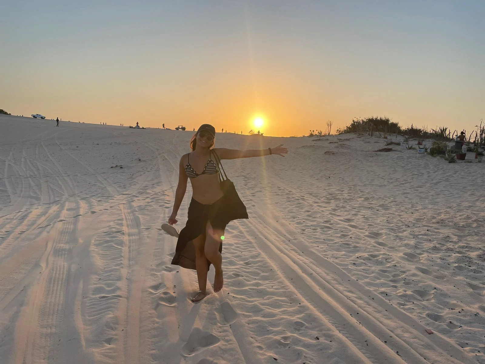 Woman standing on a sandy beach at sunset with warm orange light and visible tire tracks in the sand.