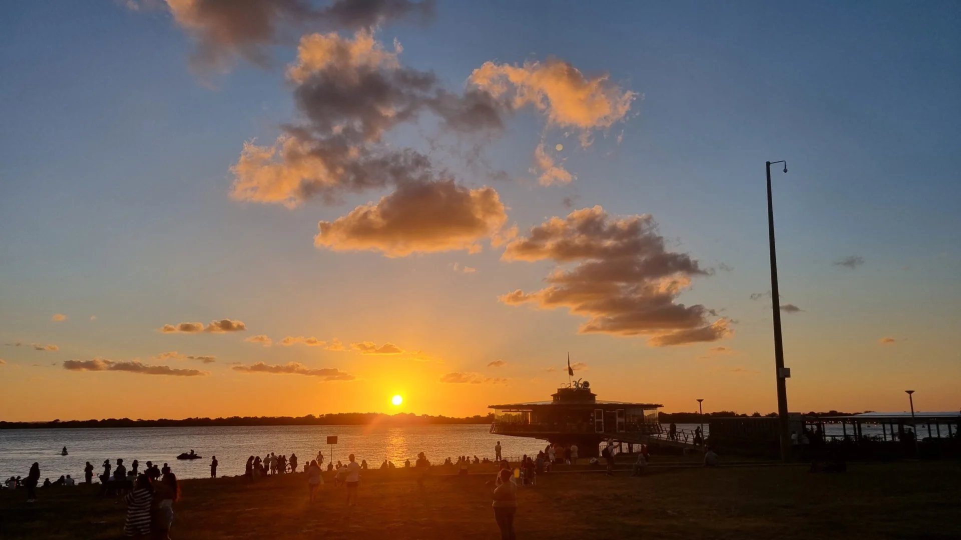 Sunset over the water with people gathered along the shore and a small structure nearby.