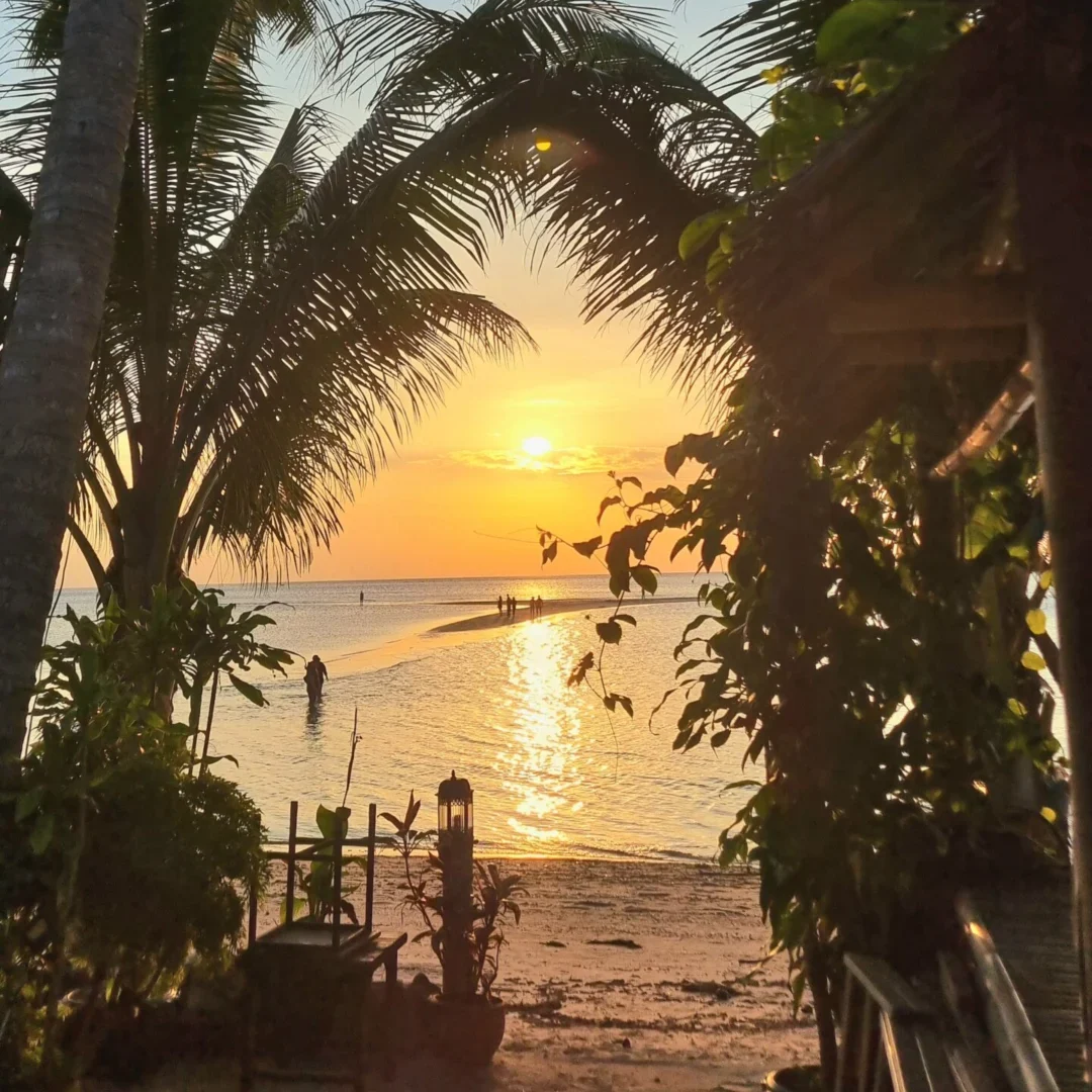 The sun sets over the sea, framed by palm trees and the dark silhouette of a bungalow at the edge of the beach.