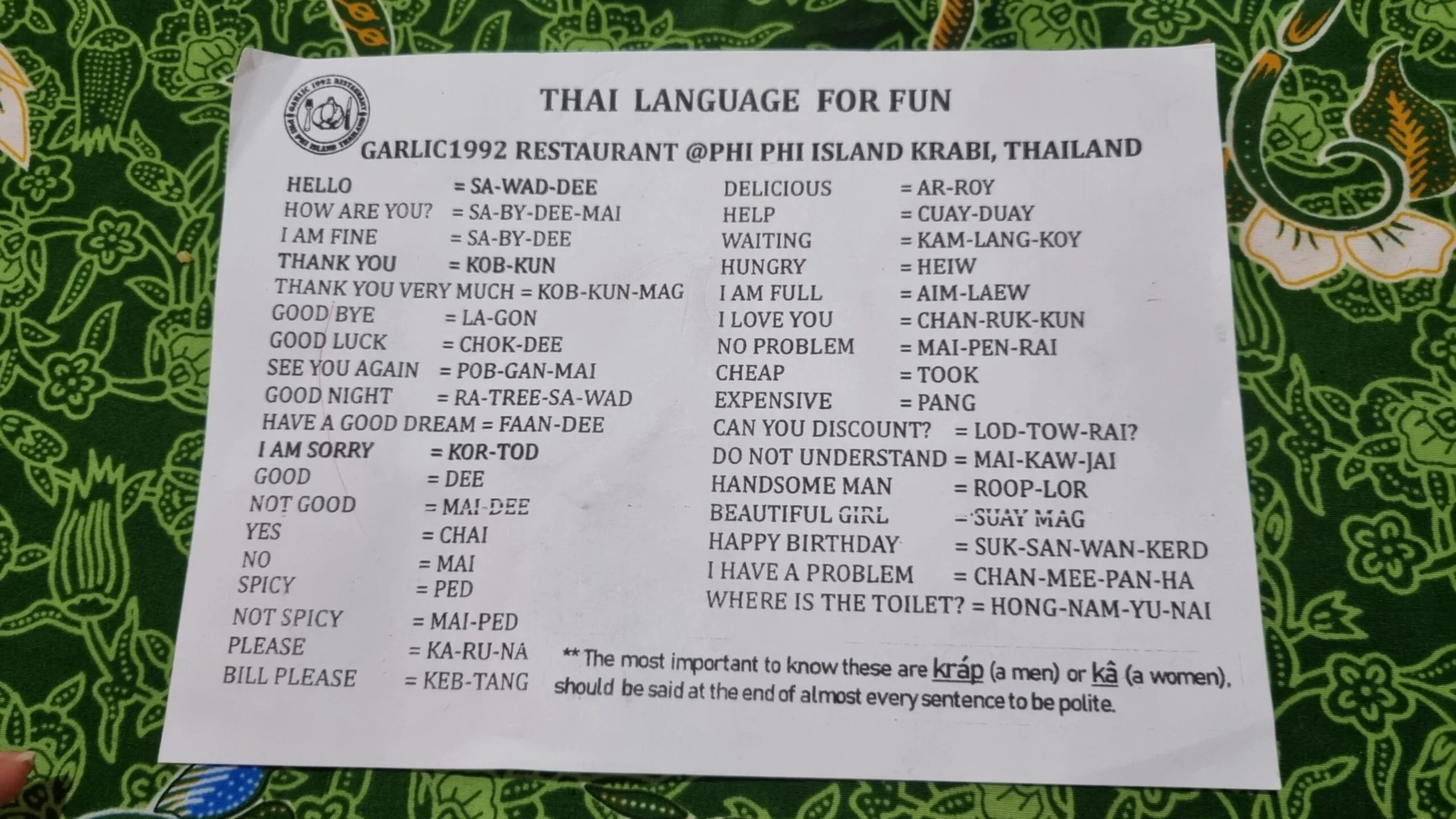 A printed Thai language cheat sheet lies on a patterned surface, showing common words and translations.