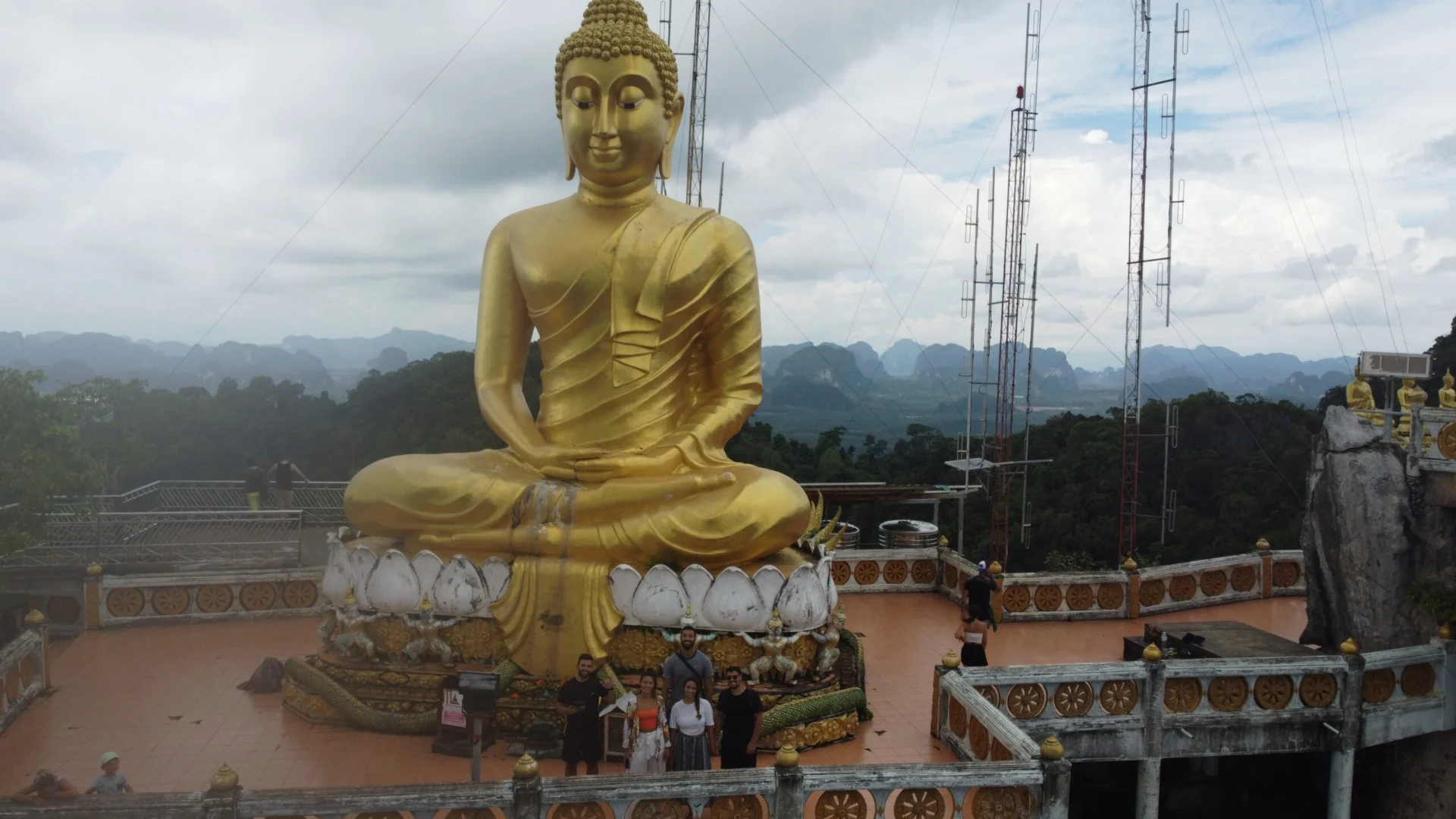 A large golden Buddha statue sits on a terrace at the top of Tiger Cave Temple with mountains in the distance.
