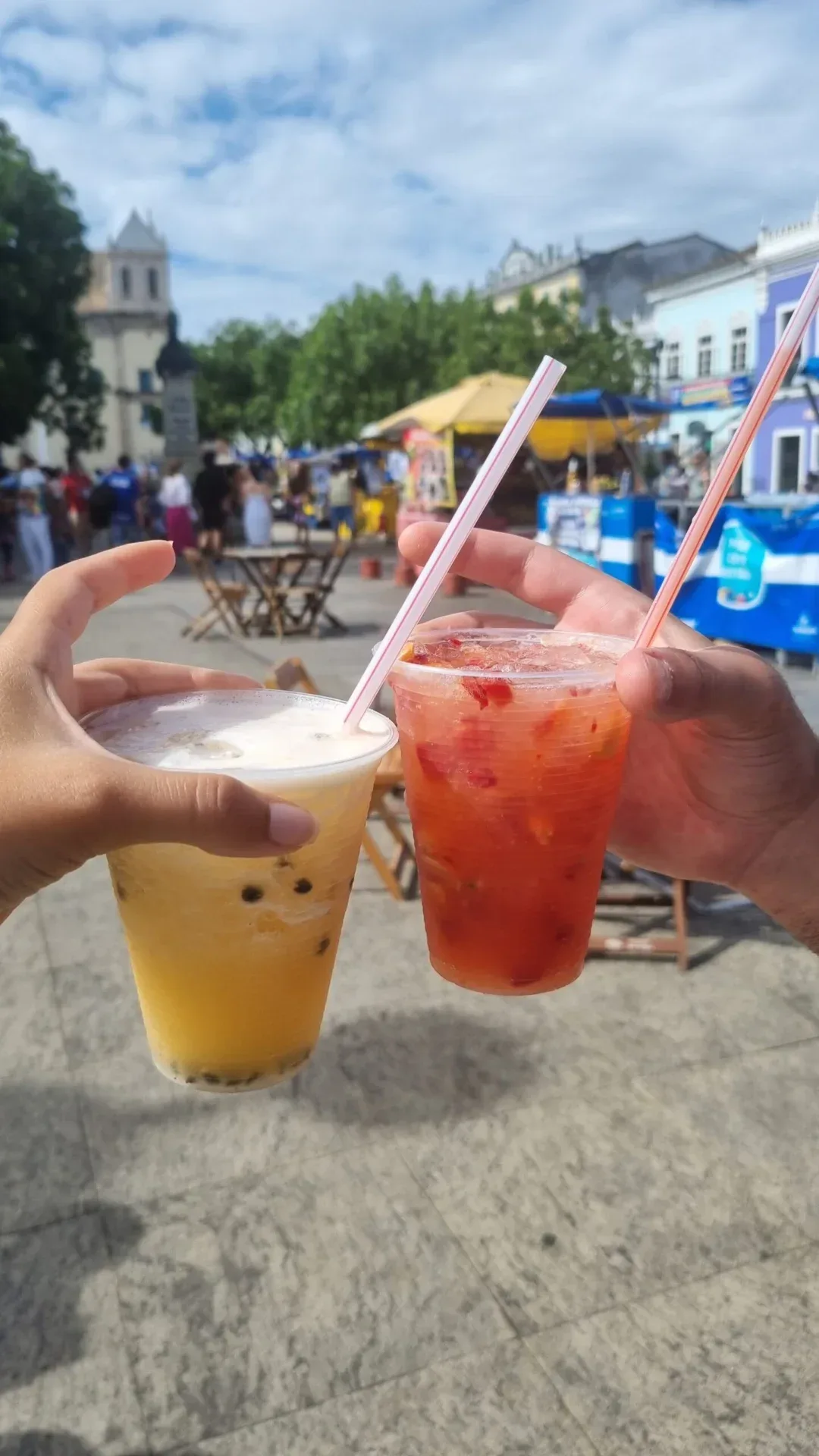 Two hands toasting with fruit drinks in plastic cups at an outdoor market