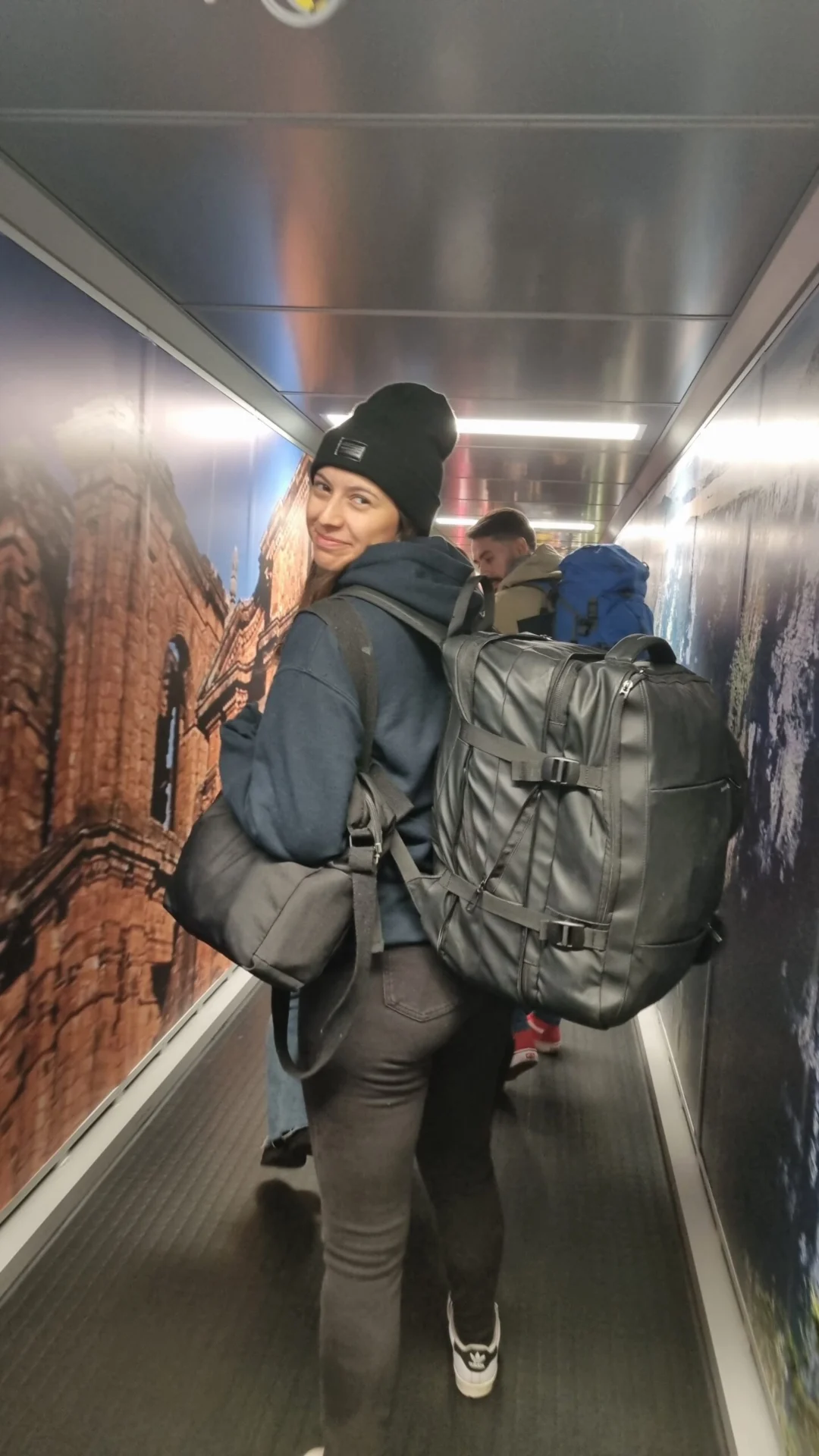 A woman with a large backpack looks back while walking through a narrow airport corridor.