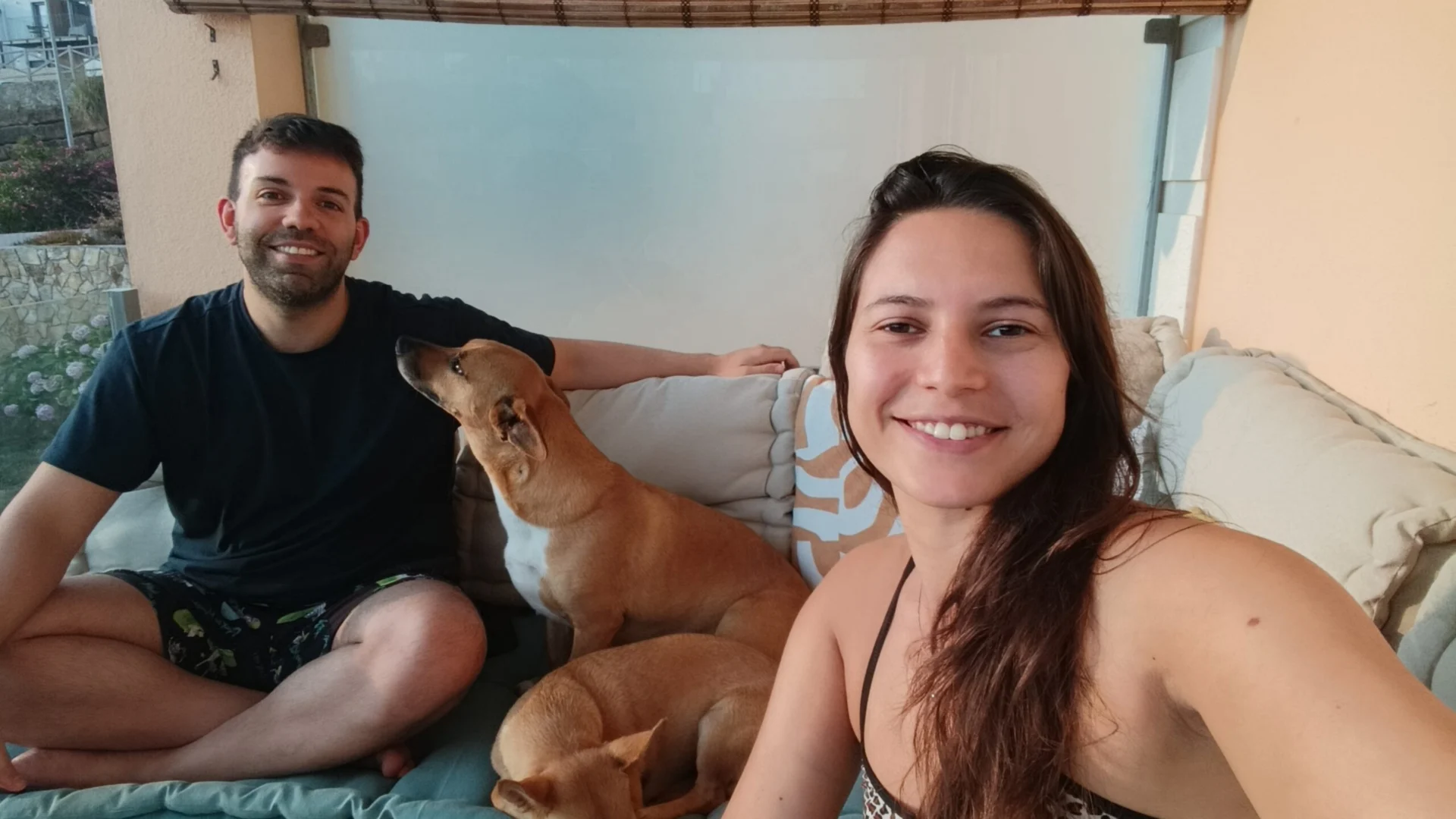 A smiling couple sits on a couch with two dogs between them.