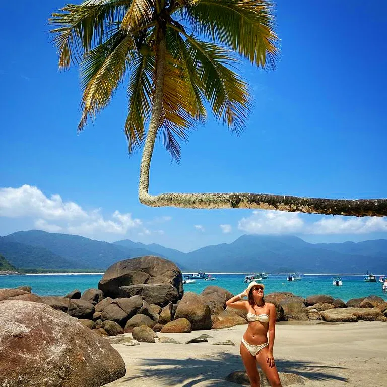 Woman in a bikini standing on a tropical beach with rocks, palm trees, blue water, and mountains.