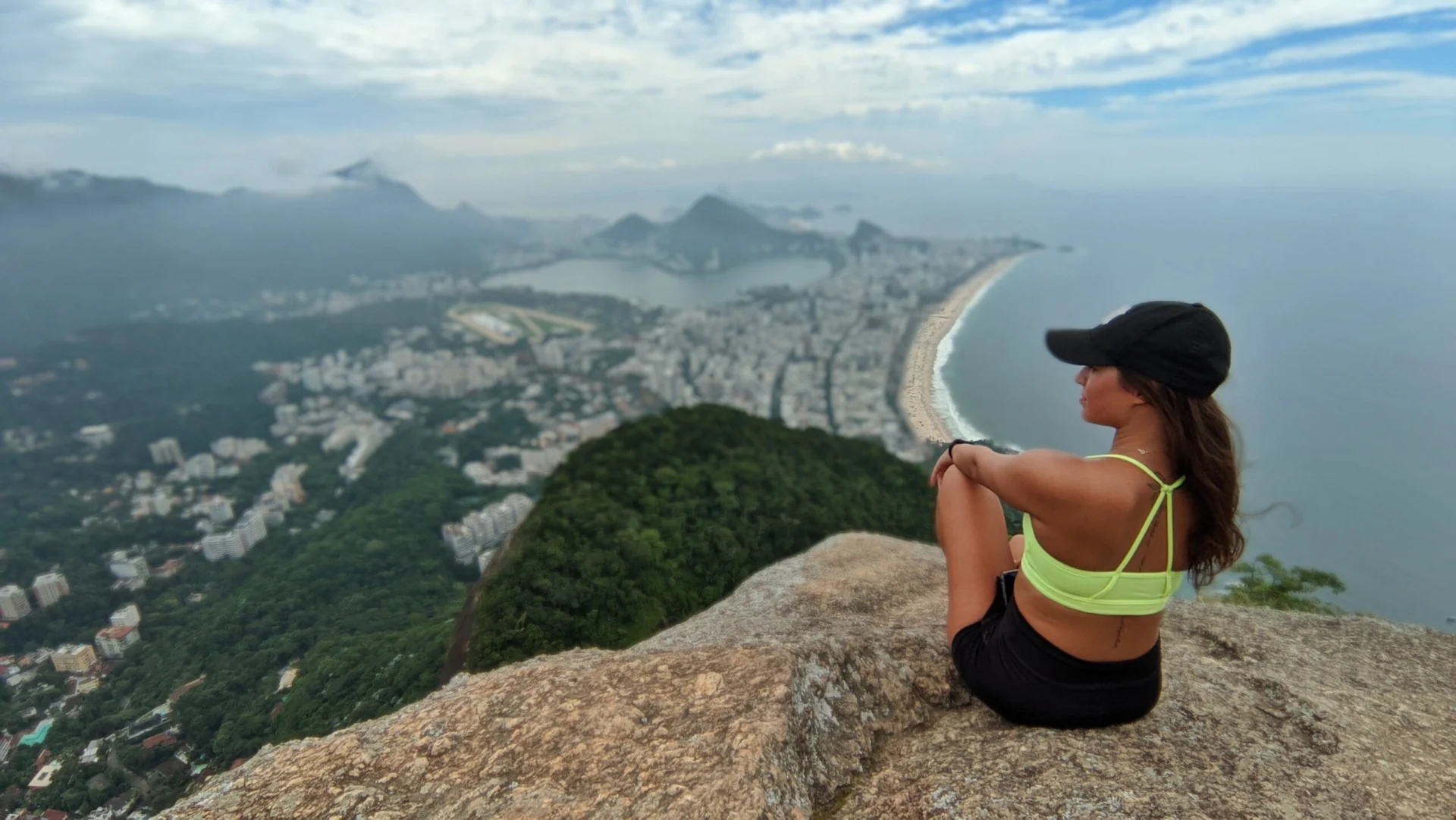 Woman sitting on a rocky viewpoint overlooking a coastal city, mountains, and a long beach.