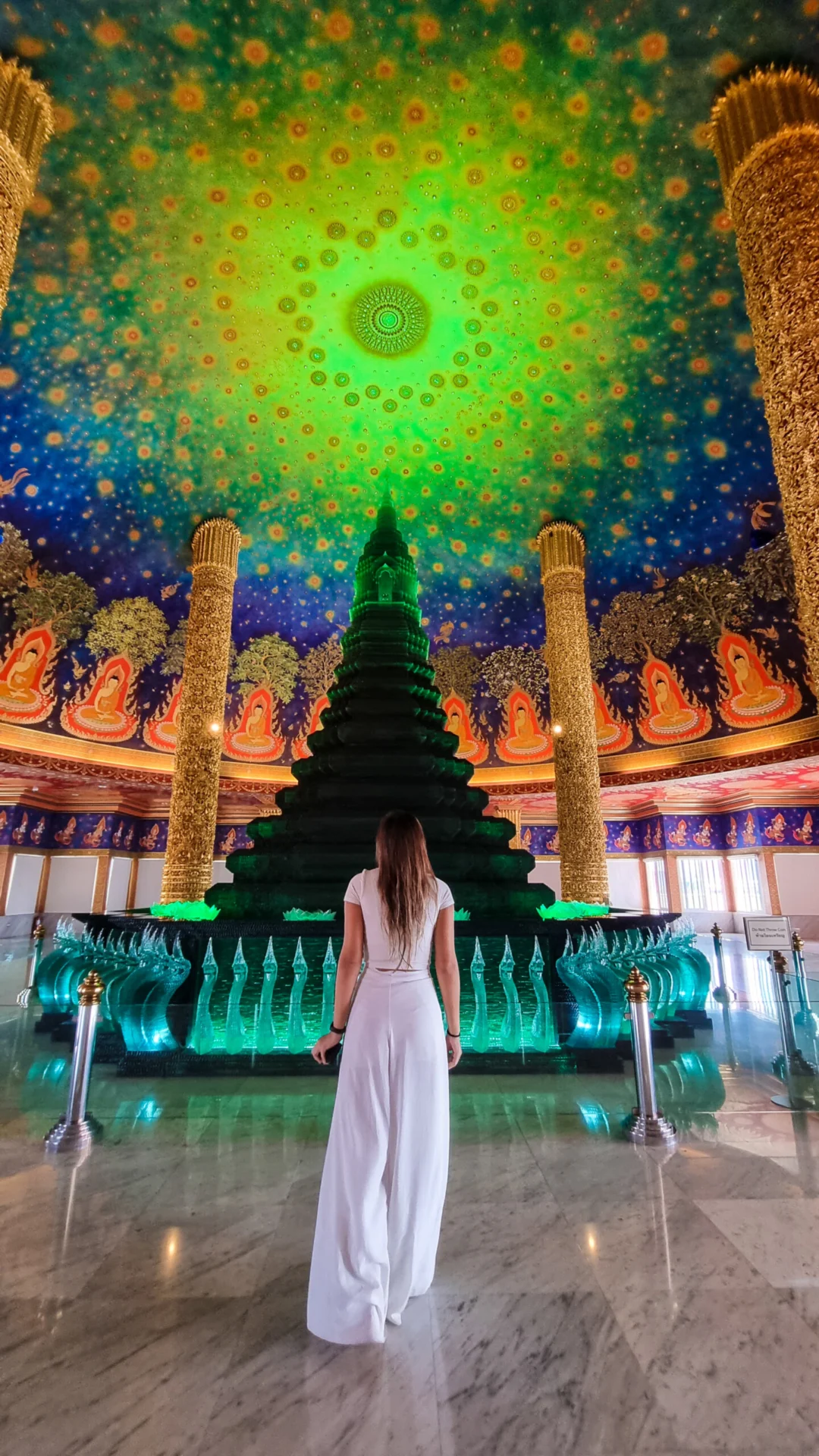 Woman in a white dress facing a glowing green stupa inside a brightly painted temple hall.