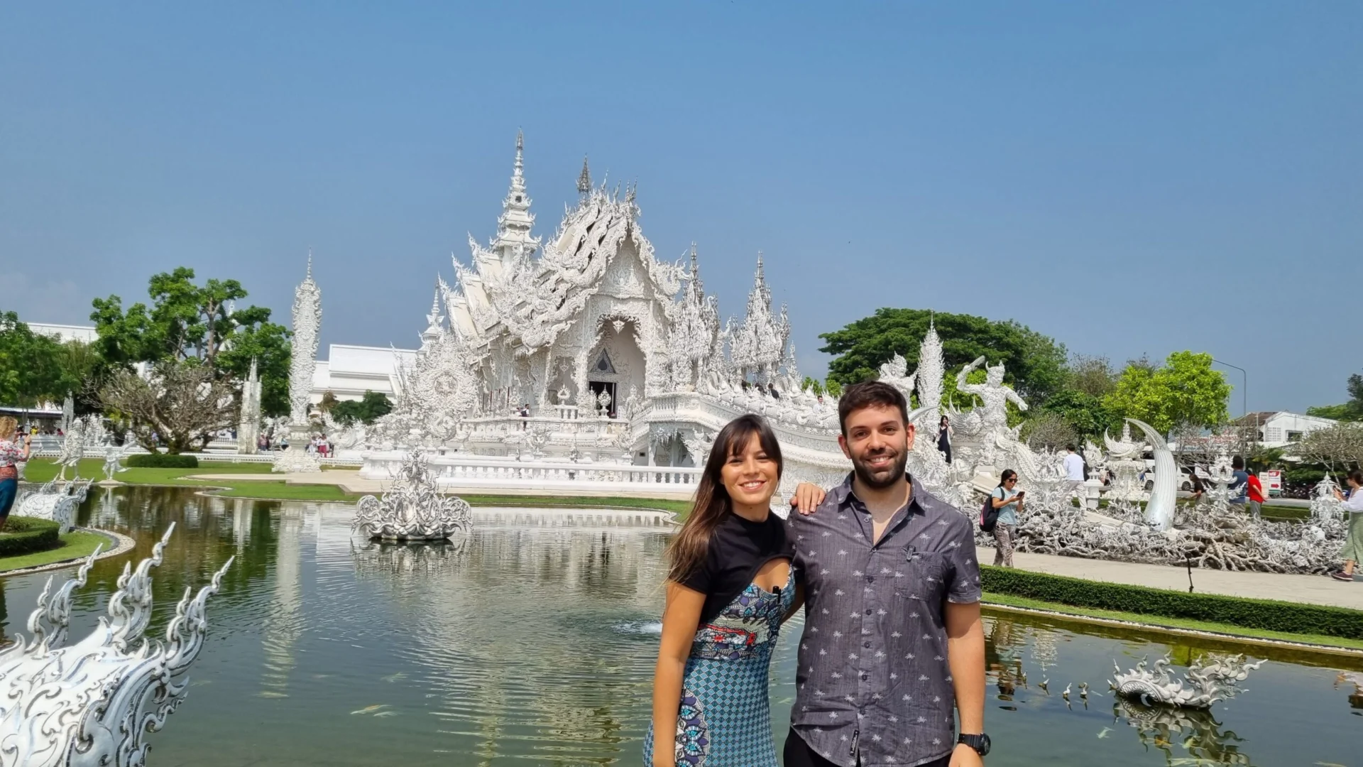A couple posing in front of the ornate white temple complex reflected in a pond.