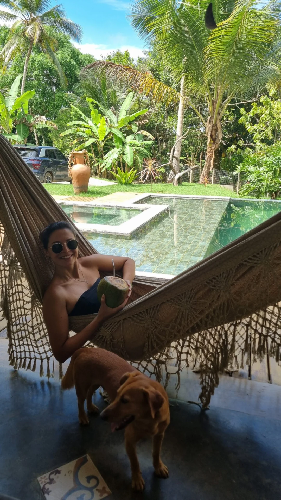 A woman relaxes in a hammock beside a pool while a small dog stands nearby.
