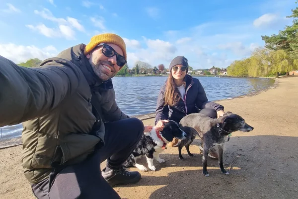 Two people crouch beside several dogs on a lakeside path during a walk.