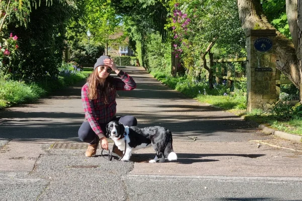 A woman crouches with a black-and-white dog on a leafy driveway.